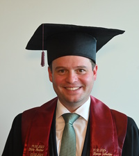 Absolvent in Hochschulrobe mit Doktorhut, rotem Stola und weißem Hemd; Name/Details auf der Stola sichtbar.