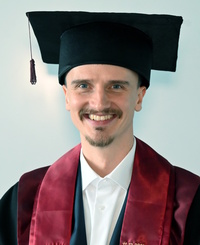 Absolvent in Graduation Cap und Robe vor hellem Hintergrund, gefasstes Gesicht.